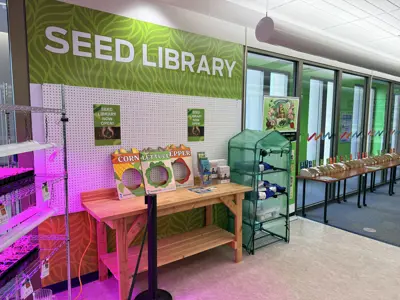 Seed Library banner over a table with seed resources, a purple glow in the left hand corner from grow lights, and a long table with seeds
